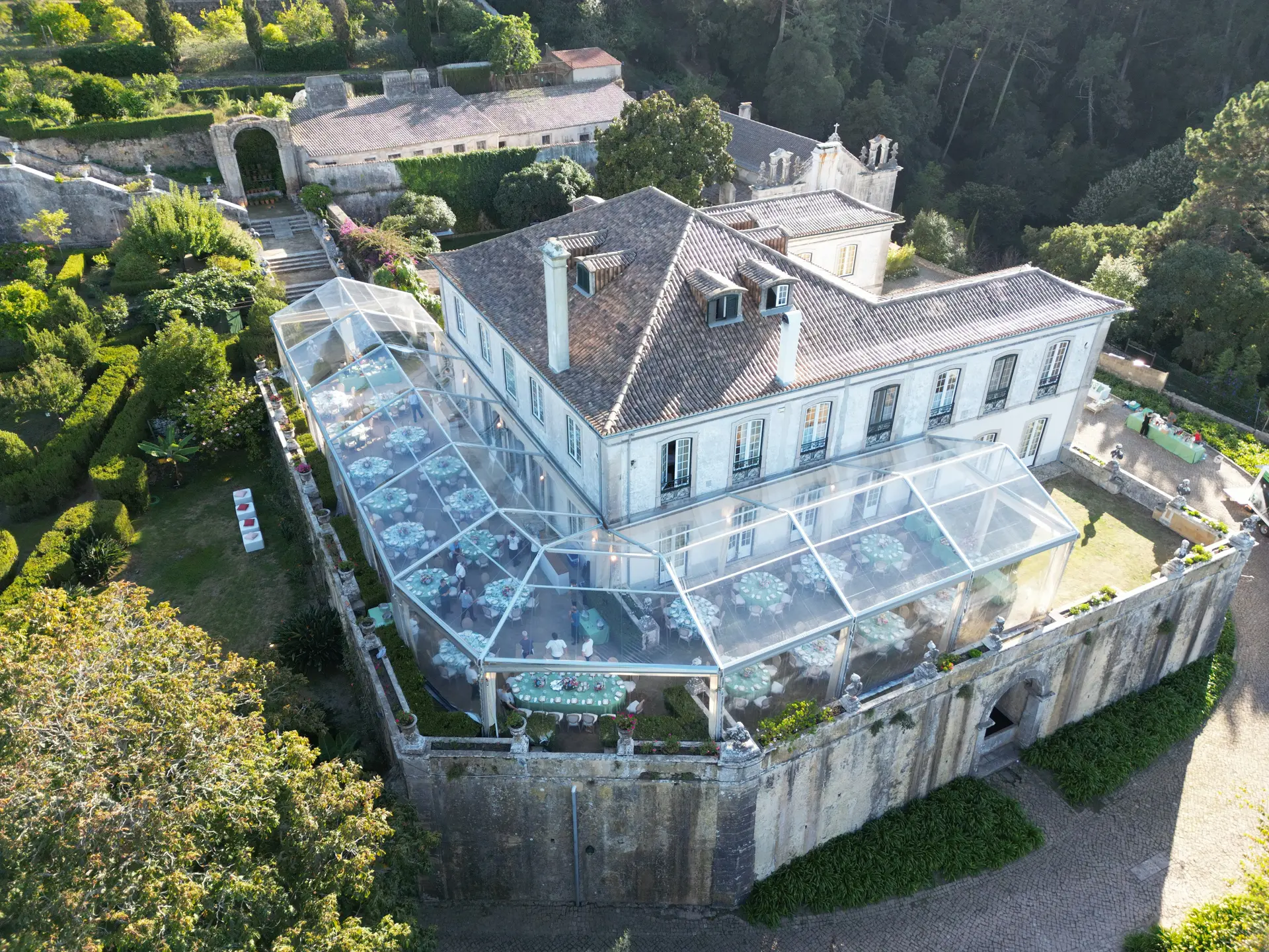 Vista aérea de uma tenda transparente de duas águas encostada a um palácio, entre jardins