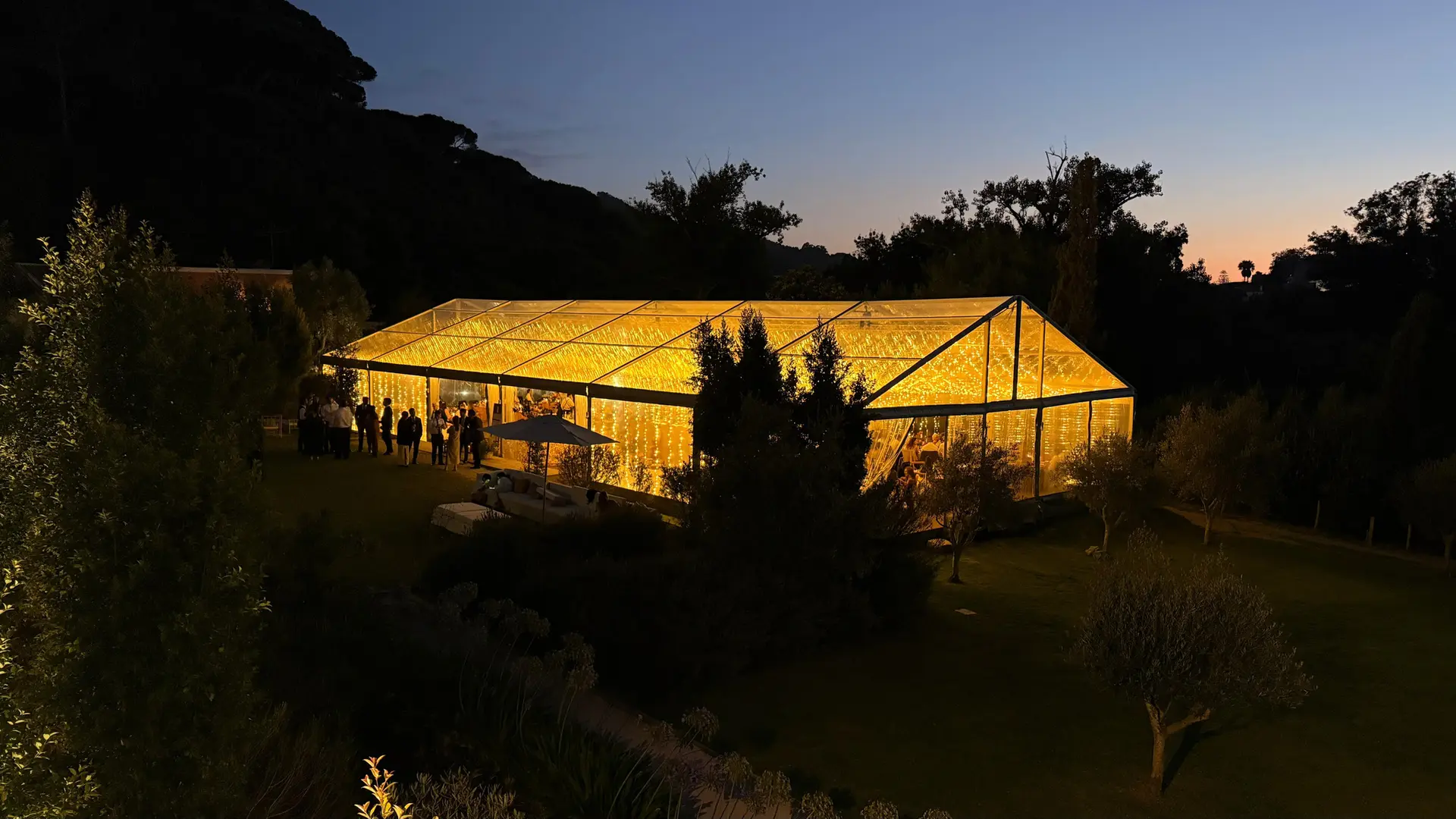 Tenda transparente acesa ao anoitecer, no meio de um jardim com árvores