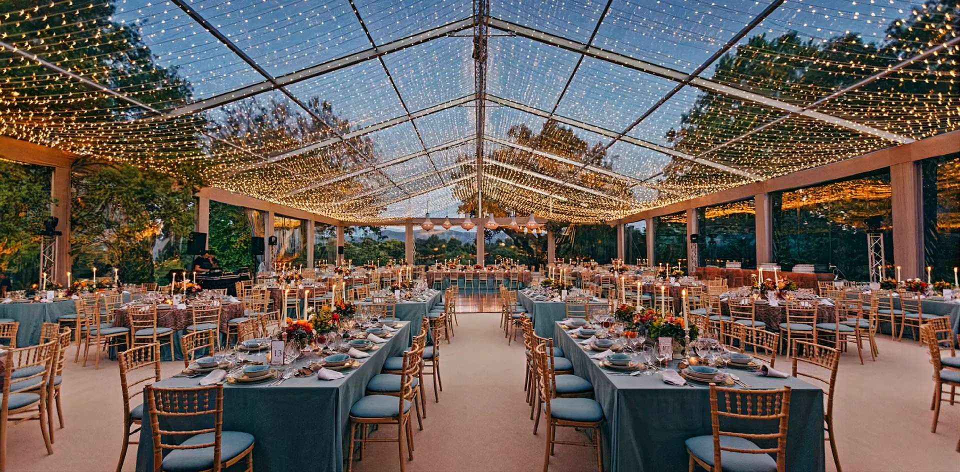 Tenda transparente com o tecto coberto de fios de luz e mesas compridas postas