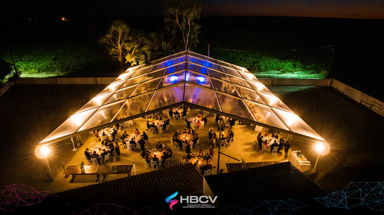 Tenda transparente vista de cima à noite, iluminada por dentro, com convidados nas mesas