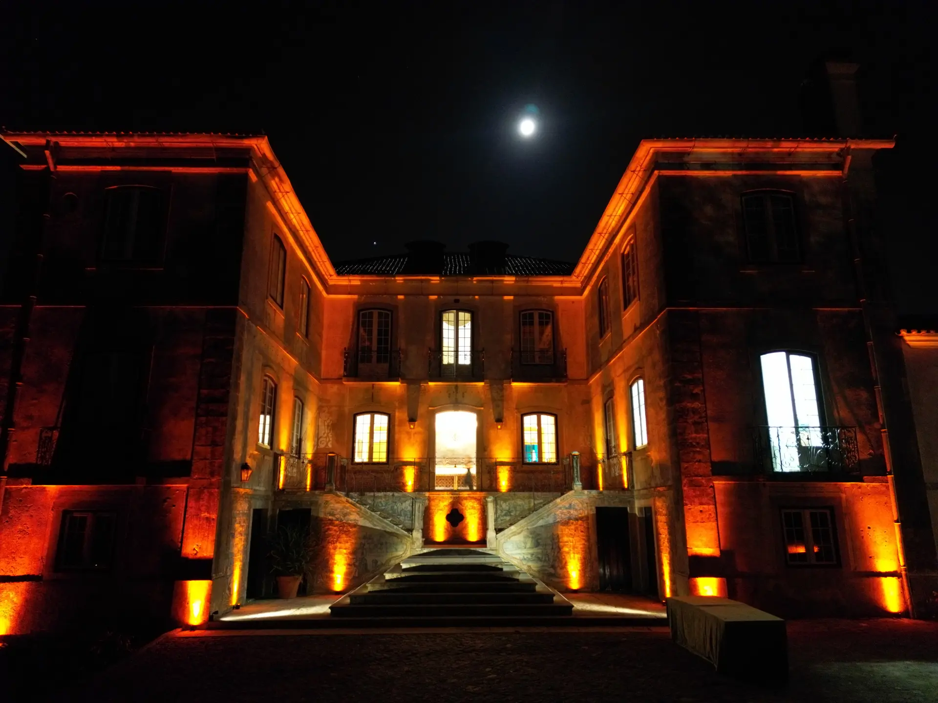 Fachada de um palácio iluminada de noite em tons quentes, com a lua por cima