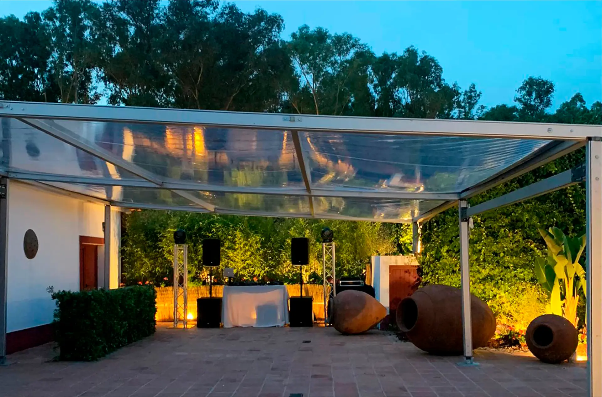 Pala transparente iluminada à noite sobre um pátio com plantas e grandes potes de barro