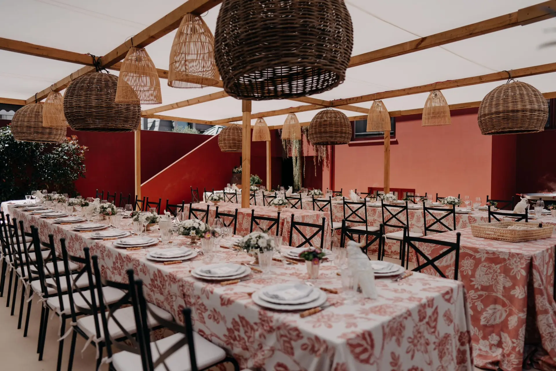 Mesas compridas com toalhas estampadas debaixo de uma pérgola com candeeiros de vime
