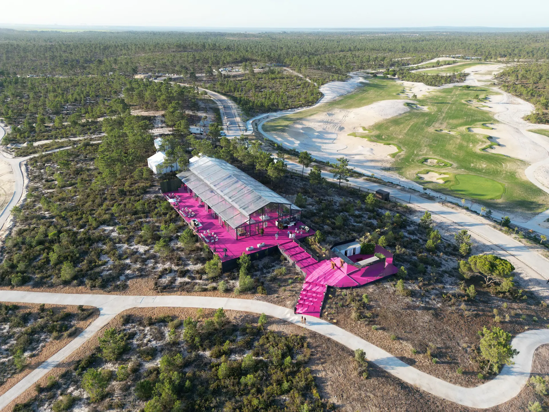 Vista aérea de uma tenda transparente de quatro águas no meio do pinhal, com passadiço cor-de-rosa