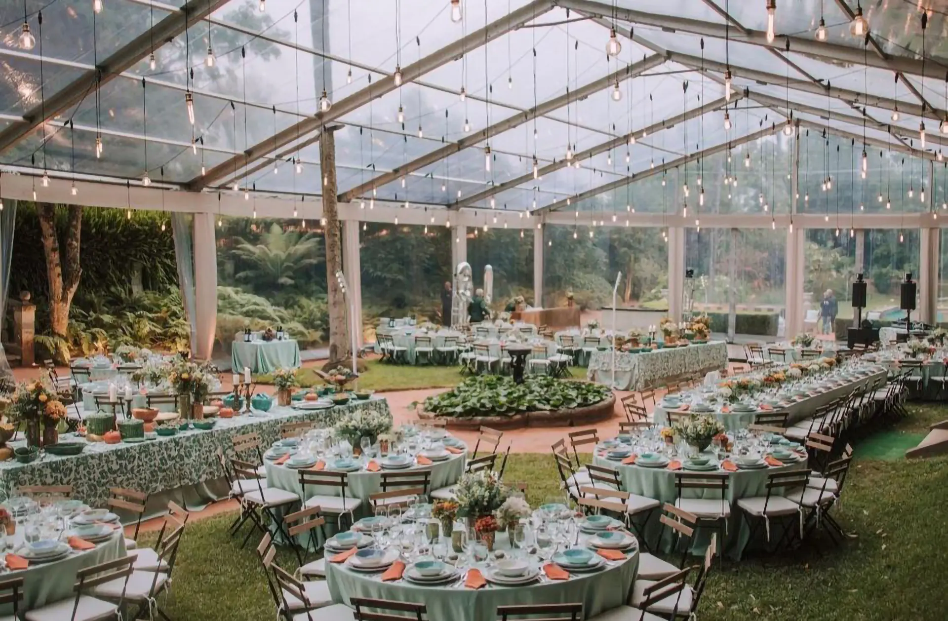 Tenda transparente montada num jardim, com mesas redondas postas para um casamento e luzes suspensas na estrutura