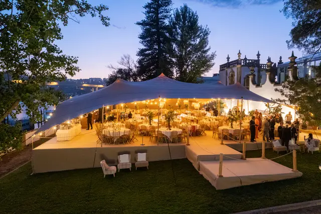 Tenda tensionada iluminada sobre mesas de jantar num relvado, com um palácio ao fundo